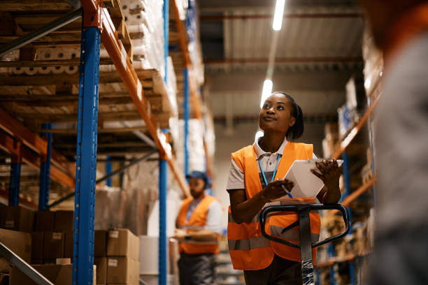 warehouss African American female worker going through inventory list while checking stock at distribution warehouse.